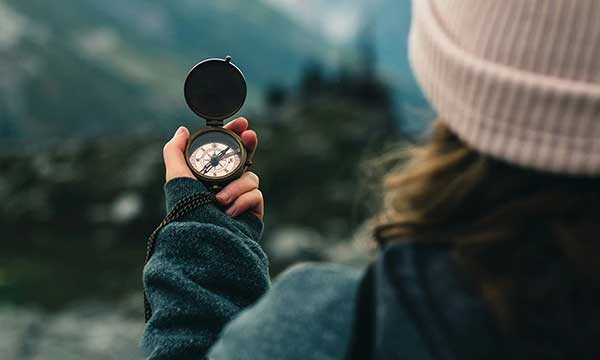 A person in a knit hat and outdoor jacket holding a brass compass up toward a blurred mountain landscape, using it to find direction on a trail.