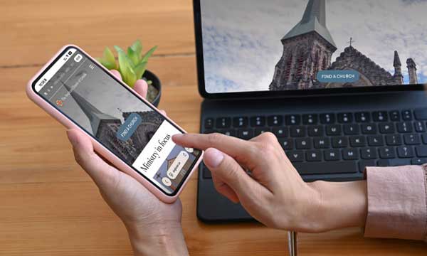 A person using a smartphone to browse the Anglican Church of Canada’s new website, with the same church-steeple page open on a laptop in the background showing a “Find a Church” button.