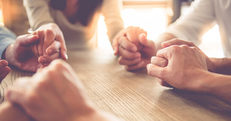 Group of hands clasped together on a wooden table, symbolizing unity and support during a gathering or meeting.