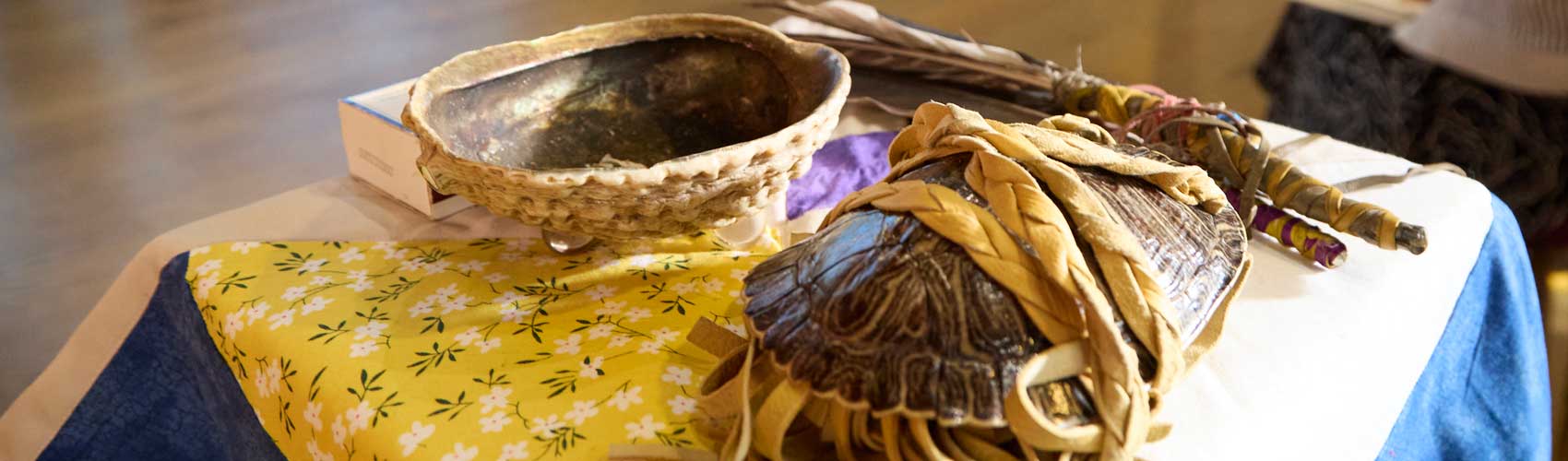 Indigenous ceremonial items arranged on a cloth-covered table, including a woven basket, a shell wrapped in braided leather, and feather bundles used for traditional smudging or prayer practices.