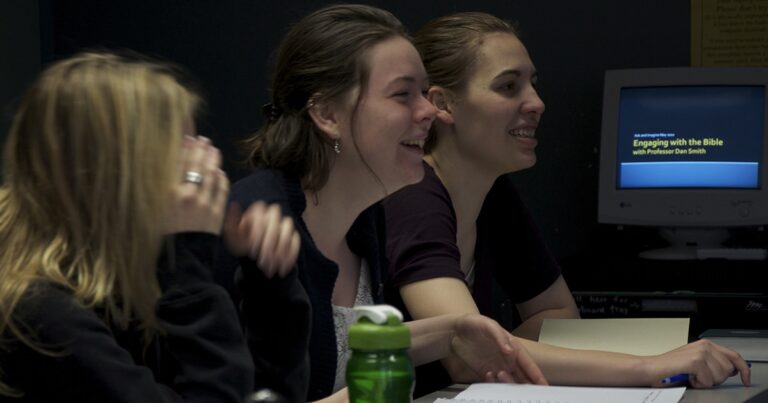 Two young women smiling and engaged in conversation, seated at a table with a water bottle in the foreground, illustrating a lively discussion or group activity.