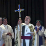 A priest in ceremonial robes holds a cross aloft during a religious service, surrounded by fellow clergy members who are applauding.