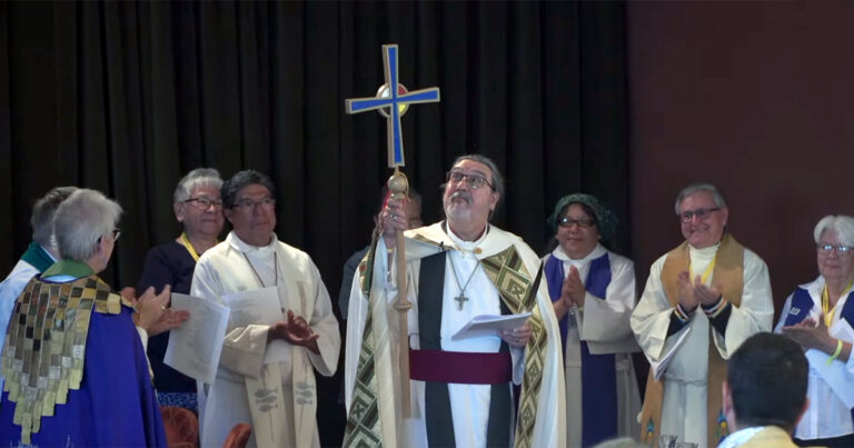 A priest in ceremonial robes holds a cross aloft during a religious service, surrounded by fellow clergy members who are applauding.