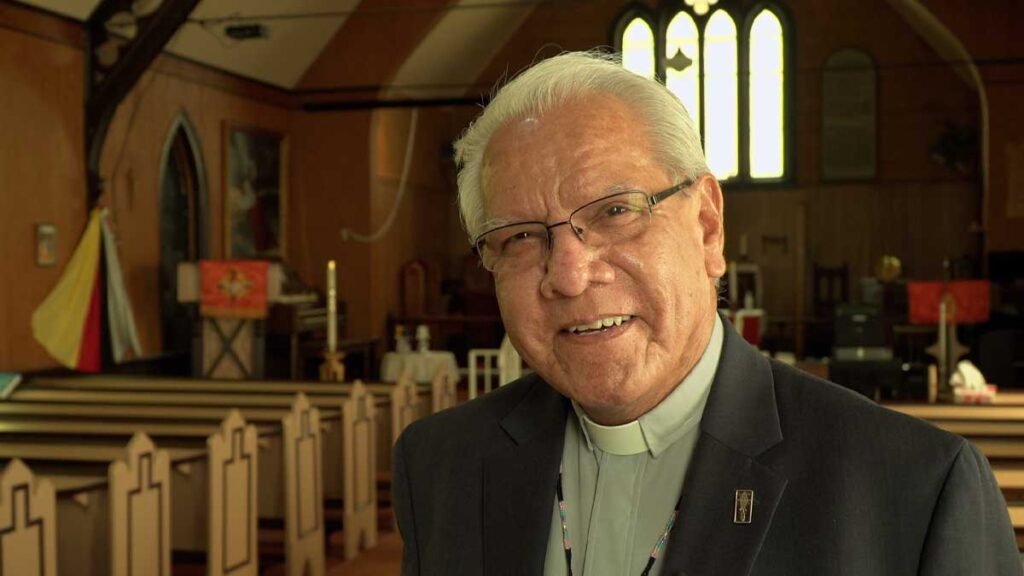 Laverne Jacobs standing inside a church, wearing glasses and a clerical collar with a dark jacket. Wooden pews and the altar are visible behind him, with warm light coming through tall stained-glass windows.