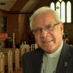 Laverne Jacobs standing inside a church, wearing glasses and a clerical collar with a dark jacket. Wooden pews and the altar are visible behind him, with warm light coming through tall stained-glass windows.