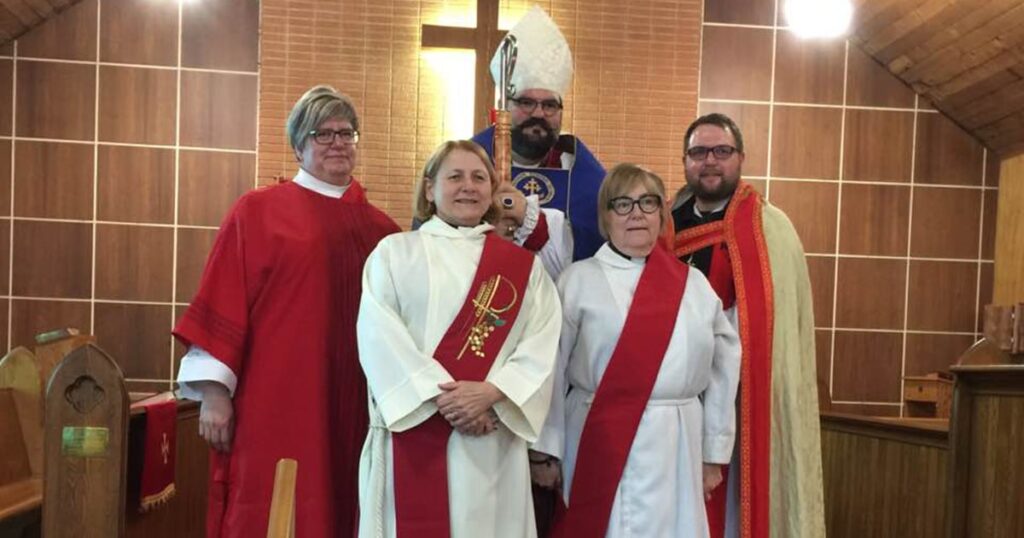 Group of clergy members standing together in a church setting, with one person in a bishop's attire holding a staff and others wearing liturgical vestments, all in front of a wooden cross backdrop.