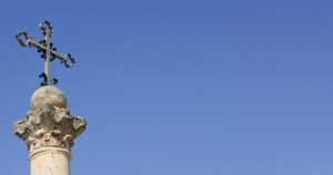 Stone column with an ornate carved capital topped by a metal cross, set against a clear blue sky.