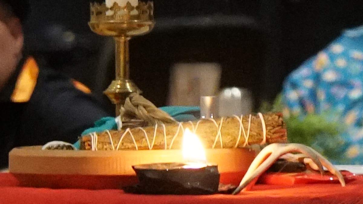 A lit candle, sage smudge, and other worship items used by the Indigenous Church, on top of a table with a red table cloth.