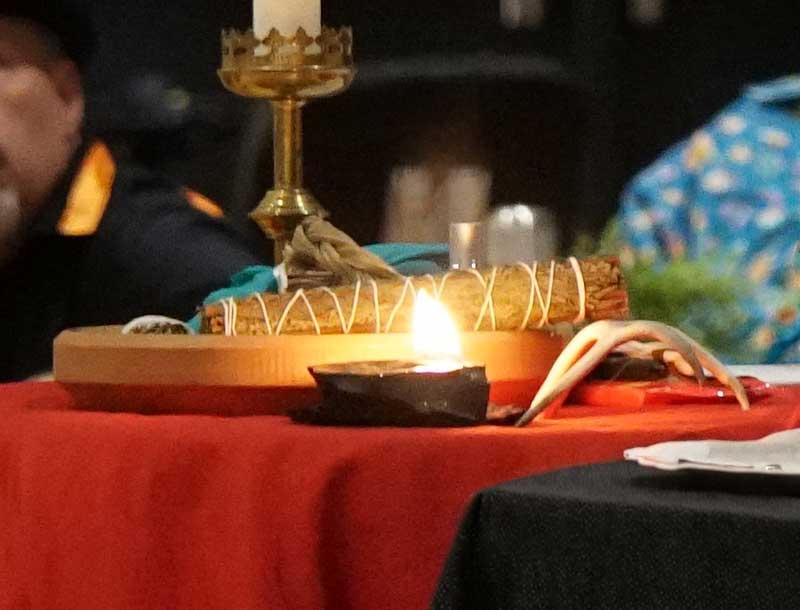 Lit candle beside ceremonial items, including a smudge bundle and antlers, arranged on a red-covered altar during a worship gathering