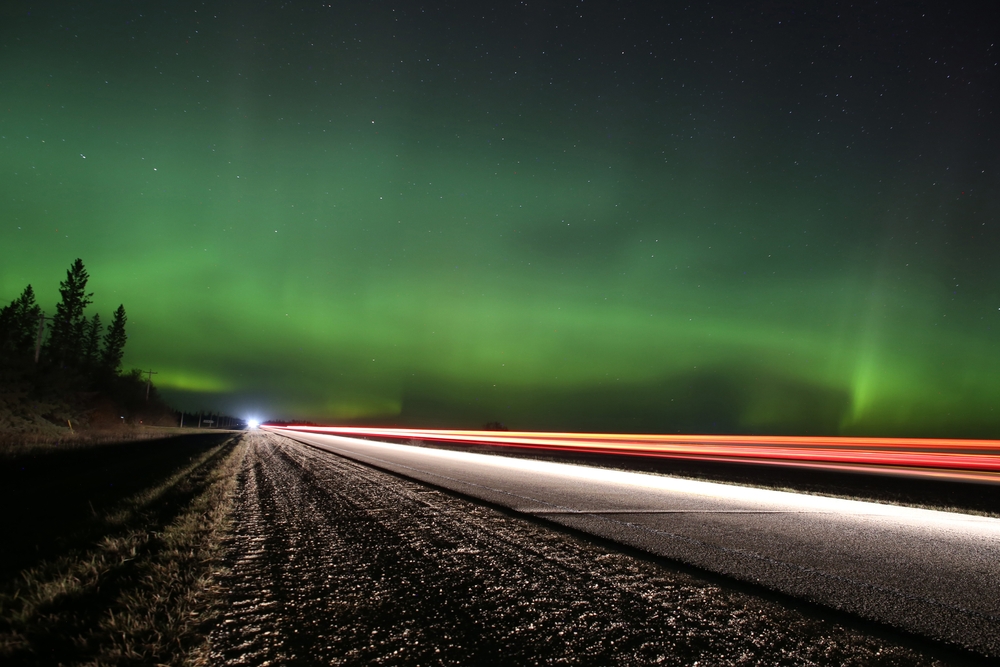 A landscape photo shows the blur of a cars lights in high speed on a roadway in a northern/remote location. The Northern Lights are seen in the sky.