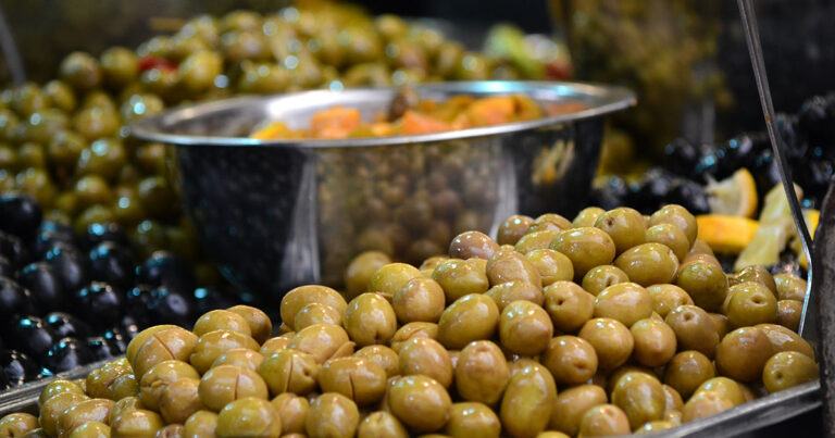 A close-up image of a pile of green olives with a stainless steel bowl of mixed pickled vegetables in the background, showcasing fresh produce at a market.