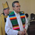 Clergy processing into a church, led by a priest wearing a white robe and a multicoloured stole, followed by bishops in liturgical vestments; parishioners look on from the pews.