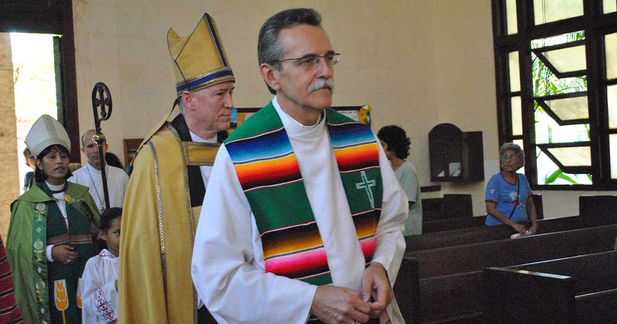 Clergy processing into a church, led by a priest wearing a white robe and a multicoloured stole, followed by bishops in liturgical vestments; parishioners look on from the pews.