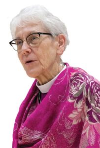 Archbishop Linda Nicholls wearing a clerical collar and a bright pink patterned shawl, looking to the side against a white background.
