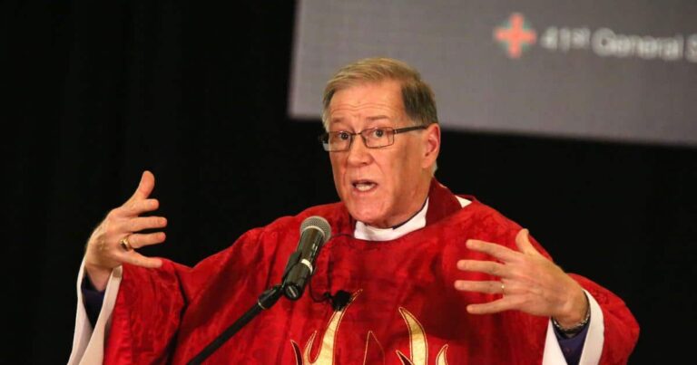 A man in a red robe gestures passionately while speaking at a podium, with a blurred background suggesting a conference setting.