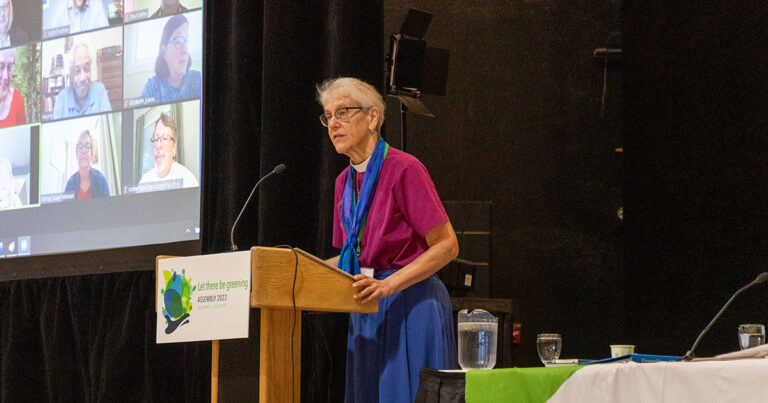 A woman with glasses speaks at a podium during a conference, wearing a pink shirt and blue skirt, with a water pitcher and green tablecloth visible.
