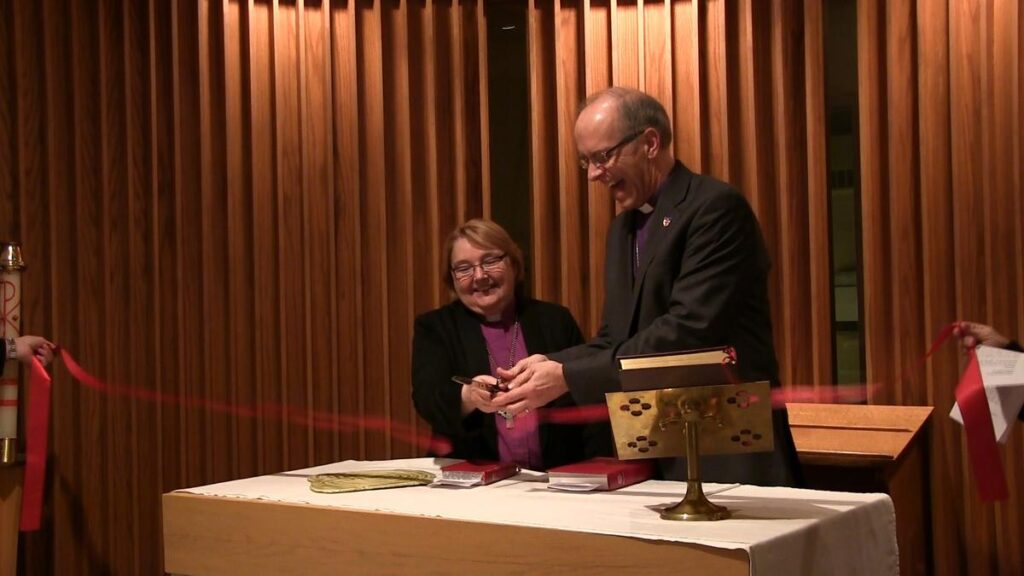 A woman and a man are smiling while cutting a ribbon together at a table, symbolizing the opening of an event or ceremony. The setting features wooden paneling in the background, adding to the formal atmosphere.