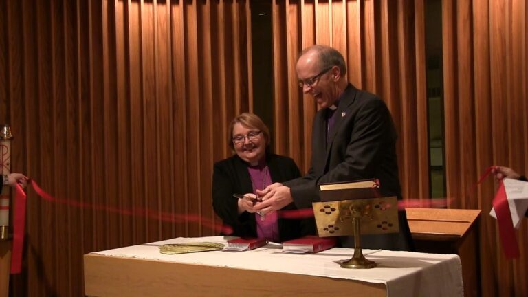A woman and a man are smiling while cutting a ribbon together at a table, symbolizing the opening of an event or ceremony. The setting features wooden paneling in the background, adding to the formal atmosphere.