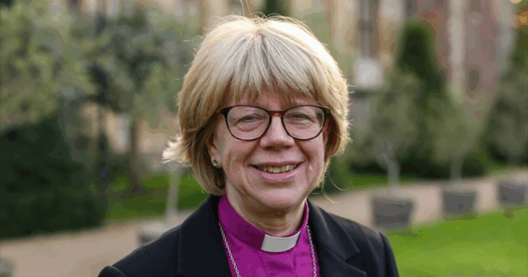 Portrait of a smiling woman with short blonde hair and glasses, wearing a black coat, set against a blurred outdoor background with greenery.