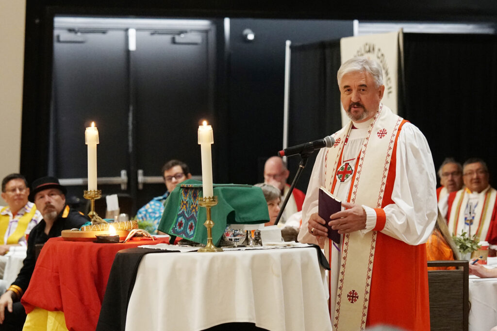 A man in religious attire speaks at a podium during a formal event, with candles and colorful ceremonial items displayed on the table in front of him.