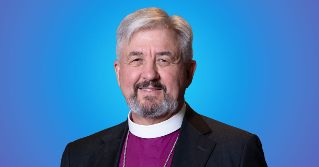Headshot of Archbishop Shane Parker, Primate of the Anglican Church of Canada, wearing clerical attire and smiling against a blue gradient background.