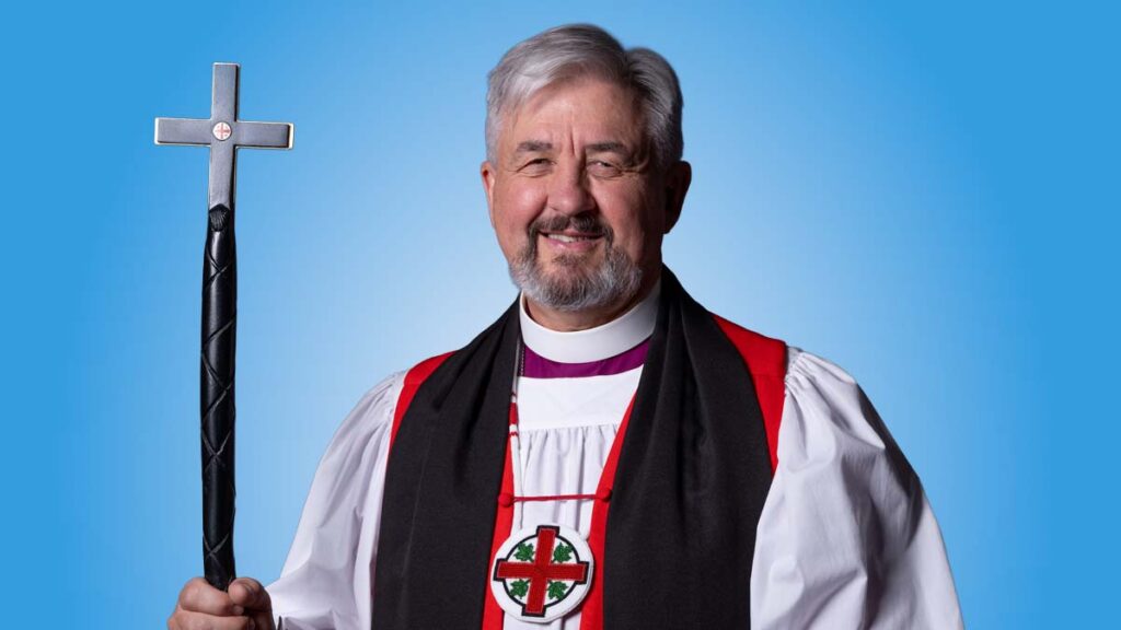 A smiling Anglican bishop wearing a white rochet with a red chimere and black stole, holding a black pastoral staff topped with a silver cross. He wears a medallion featuring the Anglican Church of Canada crest. The portrait is set against a plain light blue background.