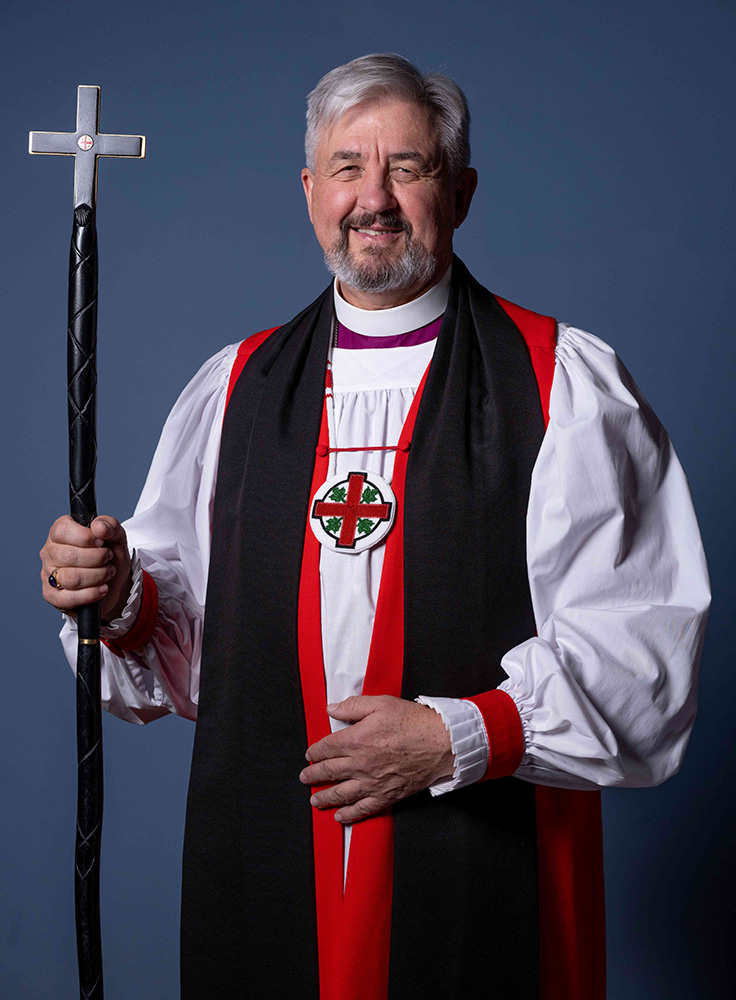 Archbishop Shane Parker, Primate of the Anglican Church of Canada, in liturgical dress holding the Primatial cross against a blue background.