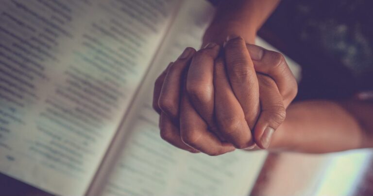Two hands clasped together in a gesture of unity, resting on an open book with visible text, symbolizing connection and shared knowledge.