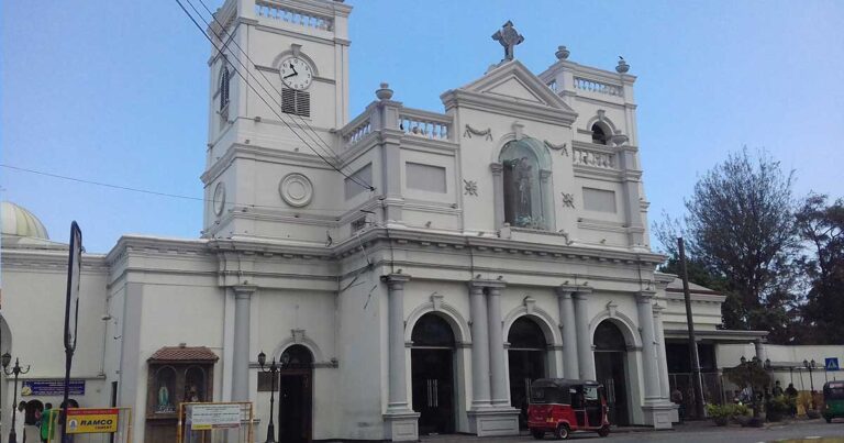Historic church building featuring a prominent clock tower, arched entrances, and decorative architectural elements, set against a clear blue sky.