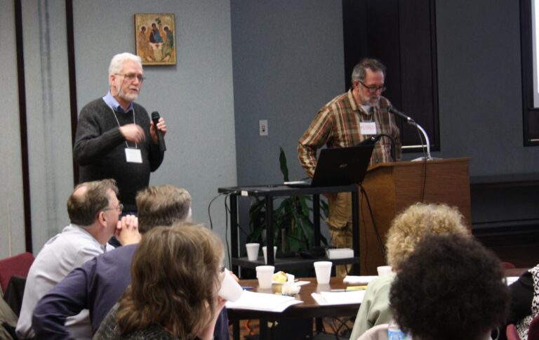 Two speakers presenting at a conference, with one using a microphone and the other standing at a podium, while an audience listens attentively.