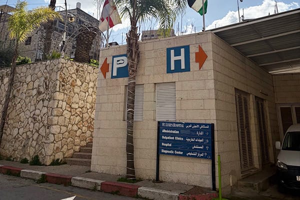 Exterior of St. Luke’s Hospital entrance with parking and hospital direction signs in English and Arabic.
