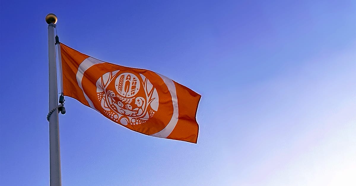 The Survivors’ Flag, bright orange with a white circular Indigenous design at the center, waves from a flagpole against a clear blue sky. The flag symbolizes remembrance, resilience, and honour for residential school survivors and victims in Canada.