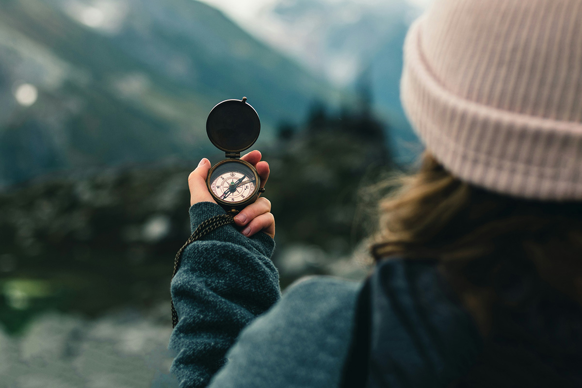 A person in a knit hat and outdoor jacket holding a brass compass up toward a blurred mountain landscape, using it to find direction on a trail.