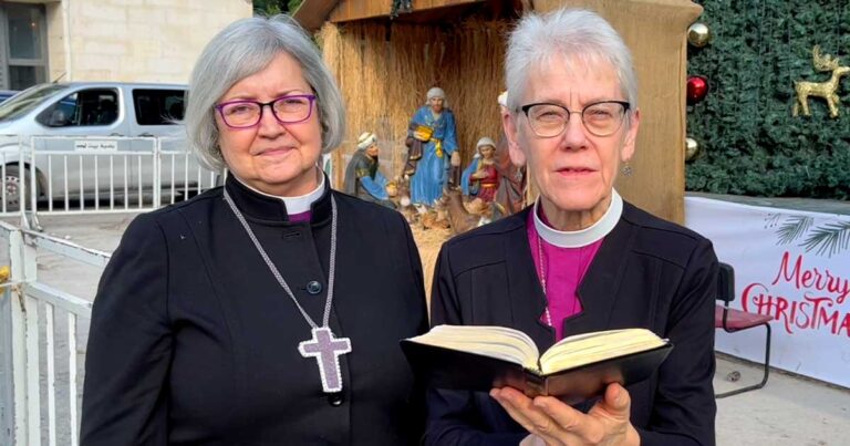 Two clergywomen holding a book, standing in front of a nativity scene. One is wearing glasses and a cross necklace, while the other has short gray hair and a similar cross. The background features figures representing the nativity.